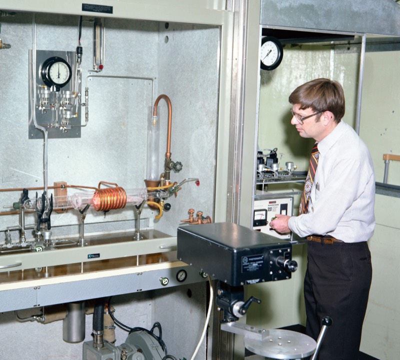 A man in a shirt and tie stands at a laboratory workstation with one hand on the controls of an instrument panel. In front of him, inside a metal-framed enclosure fitted with dial gauges, a horizontal glass tube is wrapped in a copper coil, glowing orange inside.