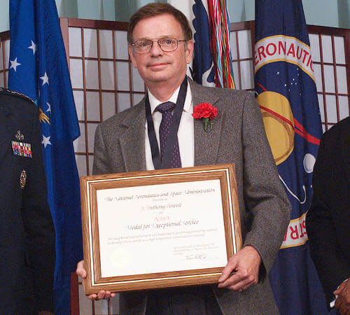 J. Anthony Powell holds a framed certificate that reads: The National Aeronautics and Space Administration presents to J. Anthony Powell the NASA Medal for Exceptional Service. He wears a grey jacket with a red carnation on the lapel; flags and a NASA seal stand behind him.